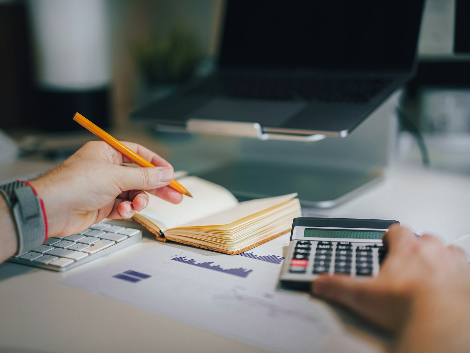 Hands holding a pencil over a notebook and working a calculator beside financial charts